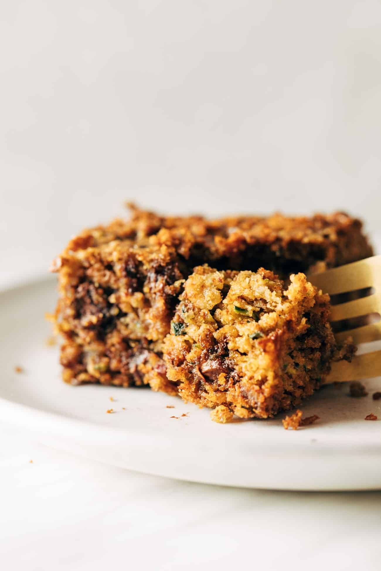 Almond butter chocolate chip zucchini bar on a white plate being cut into with a fork. 